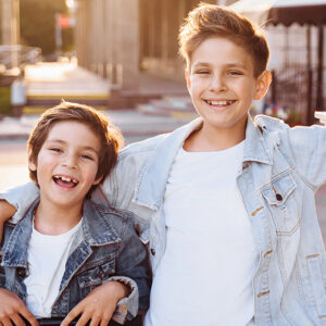 Two smiling boys wearing light denim jackets and white shirts stand outdoors with their arms around each other, enjoying a sunny day.