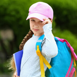 A young girl with a long braid wearing a pink cap, light blue shirt, and colorful backpack holds a blue notebook and looks at the camera, standing outdoors with green foliage in the background.